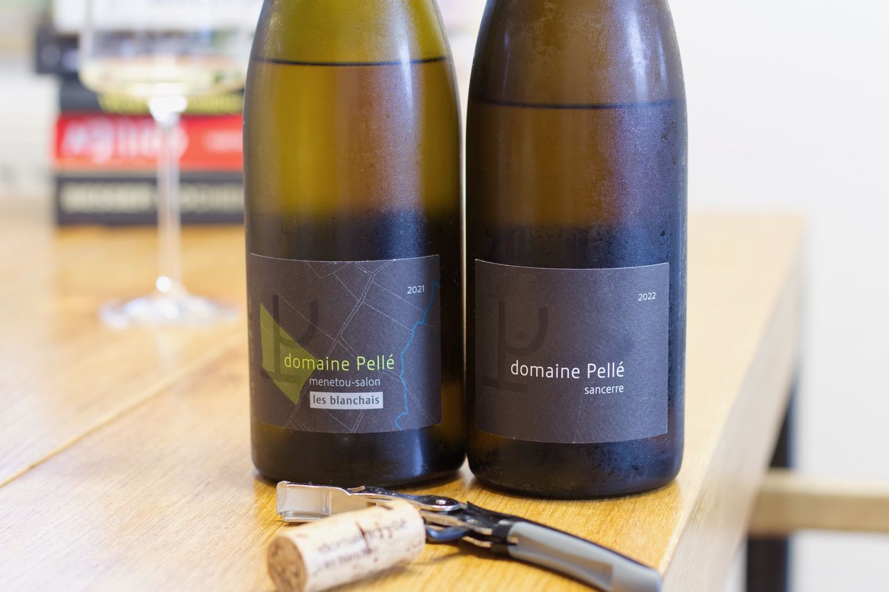 On a wooden table, there are two bottles of wine from Domaine Pellé: a Sancerre La Croix au Garde and a Menetou-Salon Les Blanchais. In the foreground, a waiter's corkscrew lies with the corks from the bottles, and in the background, a wine glass stands in front of a stack of books.
