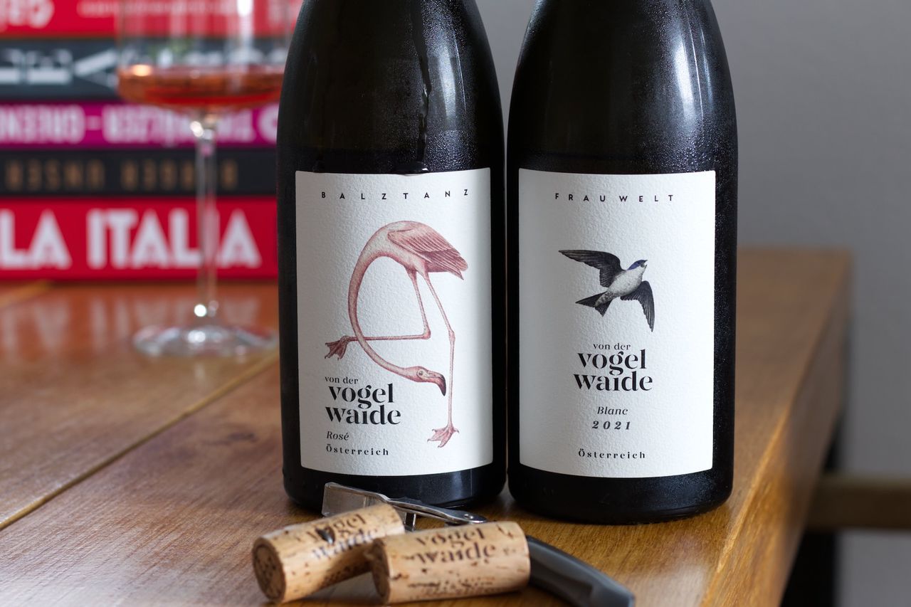 Two bottles of wine from the Von der Vogelwaide estate on a wooden table. The left bottle is a rosé named Balztanz and features a flamingo on the label. The right bottle is a white wine named Frau Welt and features a flying bird on the label. In the background, a wine glass and stacks of books can be seen. In front of the bottles are two corks and a waiter's knife.