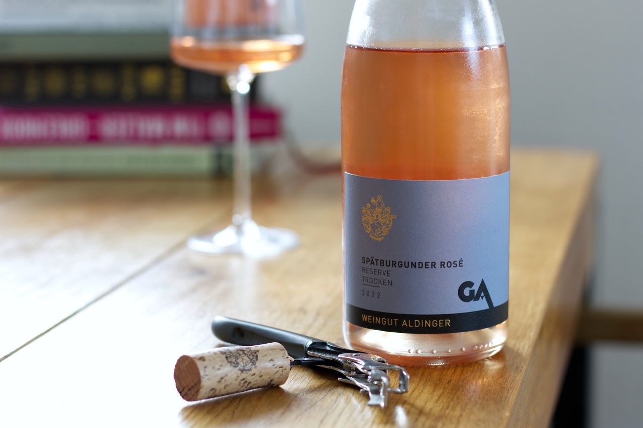 On a wooden table stands a bottle of Spätburgunder Rosé Reserve from Aldinger. In the background, a wine glass and a stack of books can be seen, while in the foreground, the cork lies on a sommelier's knife.