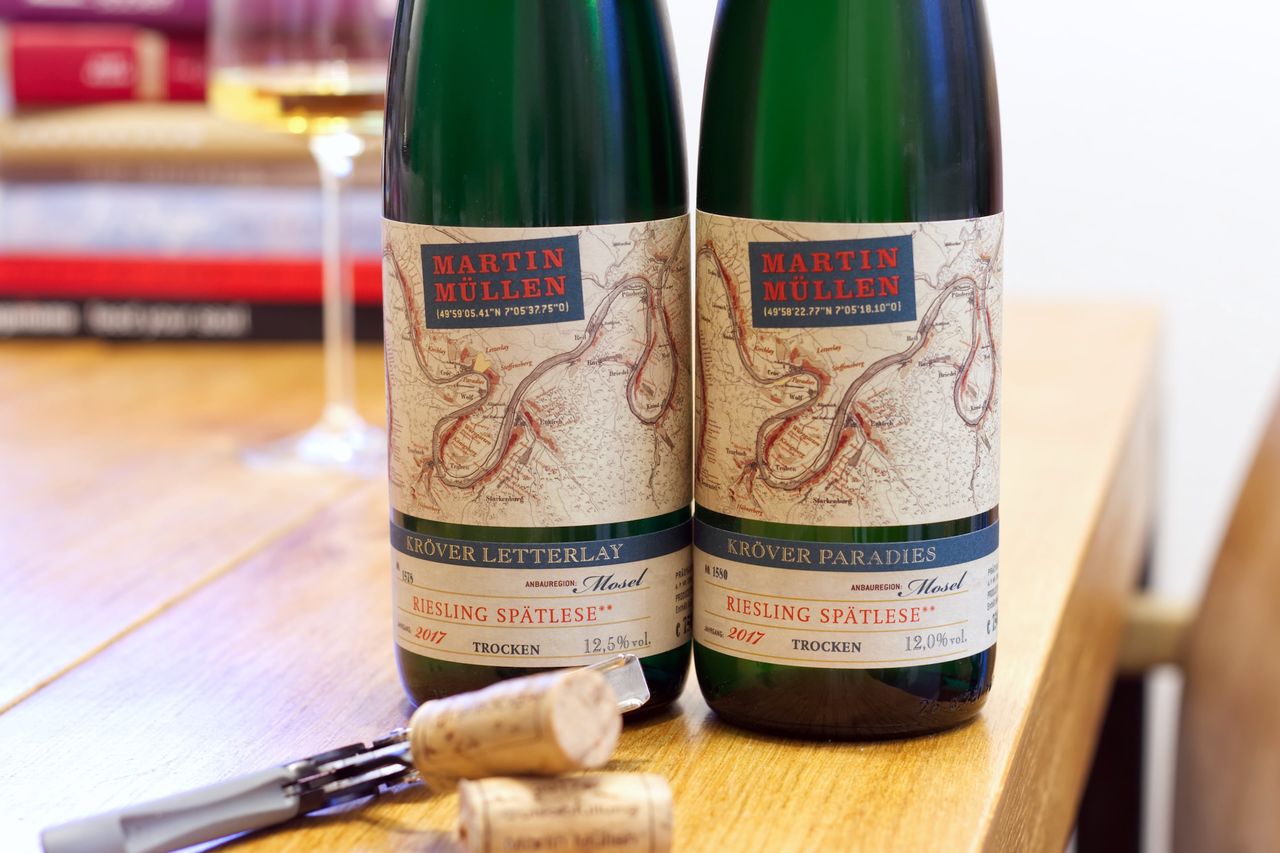On a wooden table stand two bottles of wine from Weingut Martin Müllen. One Riesling Spätlese from Kröver Paradies, one Spätlese from Kröver Letterlay. The labels show a map of the Mosel river. In the background, a wine glass and a stack of books can be seen.