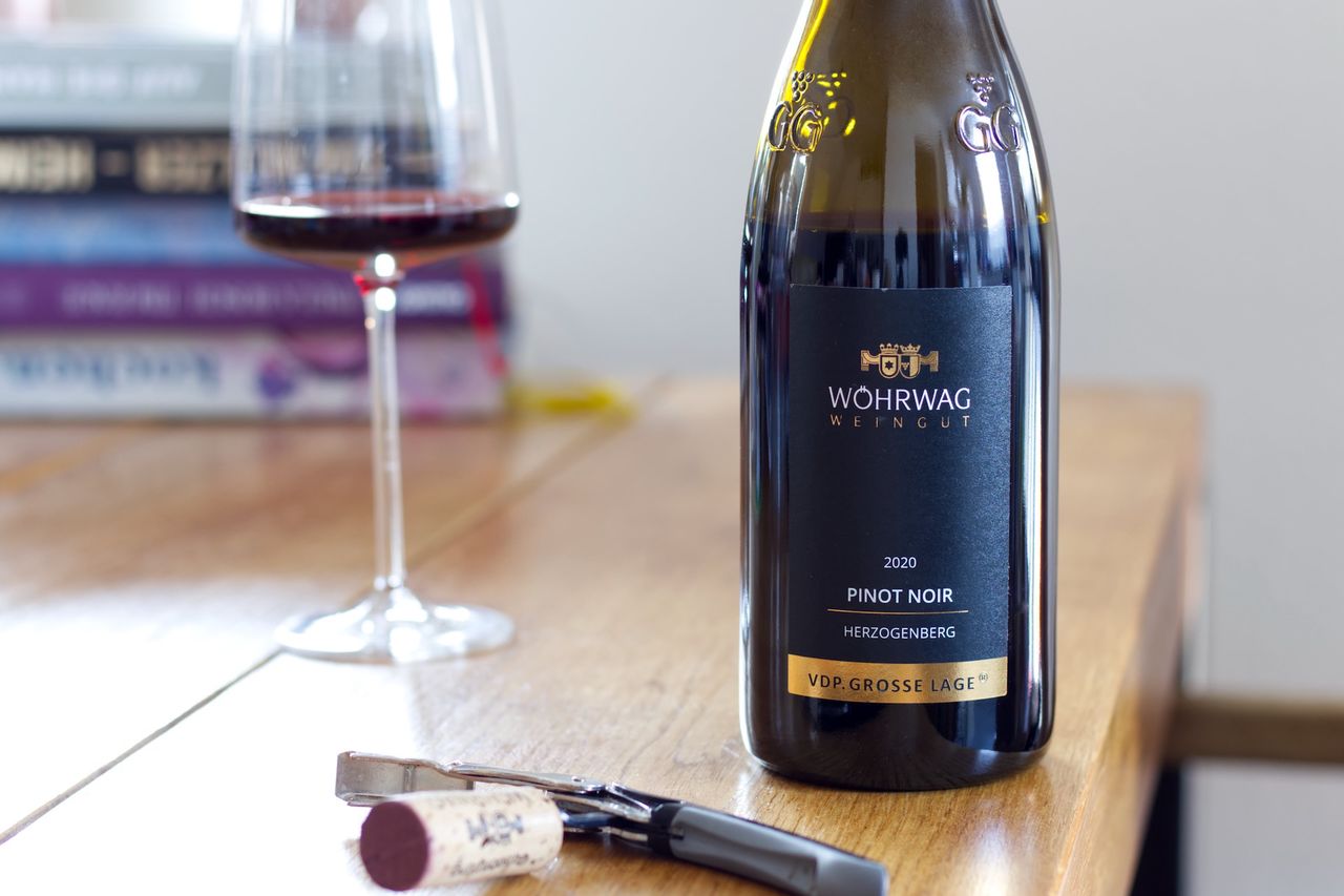 On a wooden table stands a bottle of Herzogenberg Pinot Noir from Weingut Wöhrwag. In the background, a wine glass and a stack of books can be seen, in the foreground lies the cork on the waiter's corkscrew.