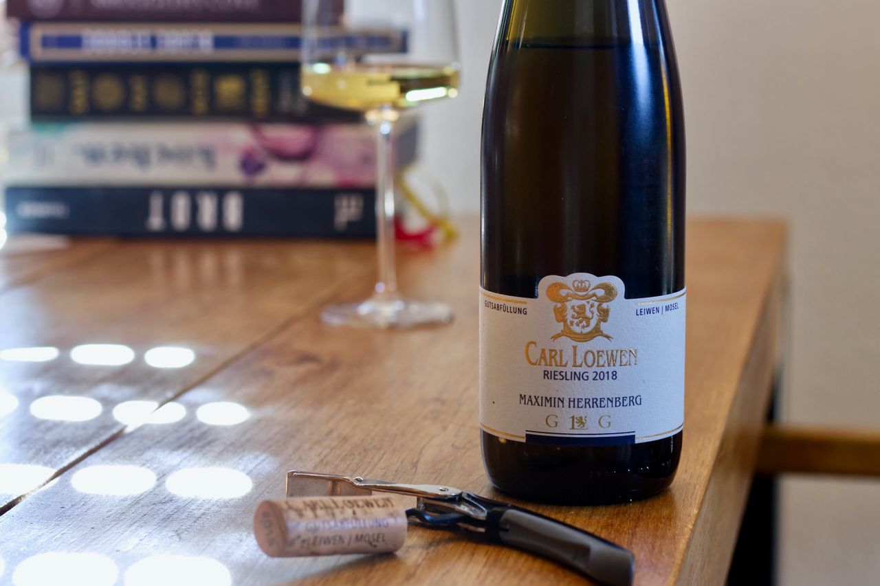 On a wooden table stands a bottle of Riesling Maximin Herrenberg from Weingut Carl Loewen from the Mosel. In the background, a wine glass and a stack of books can be seen, in the foreground, the cork lies on the waiter's corkscrew.