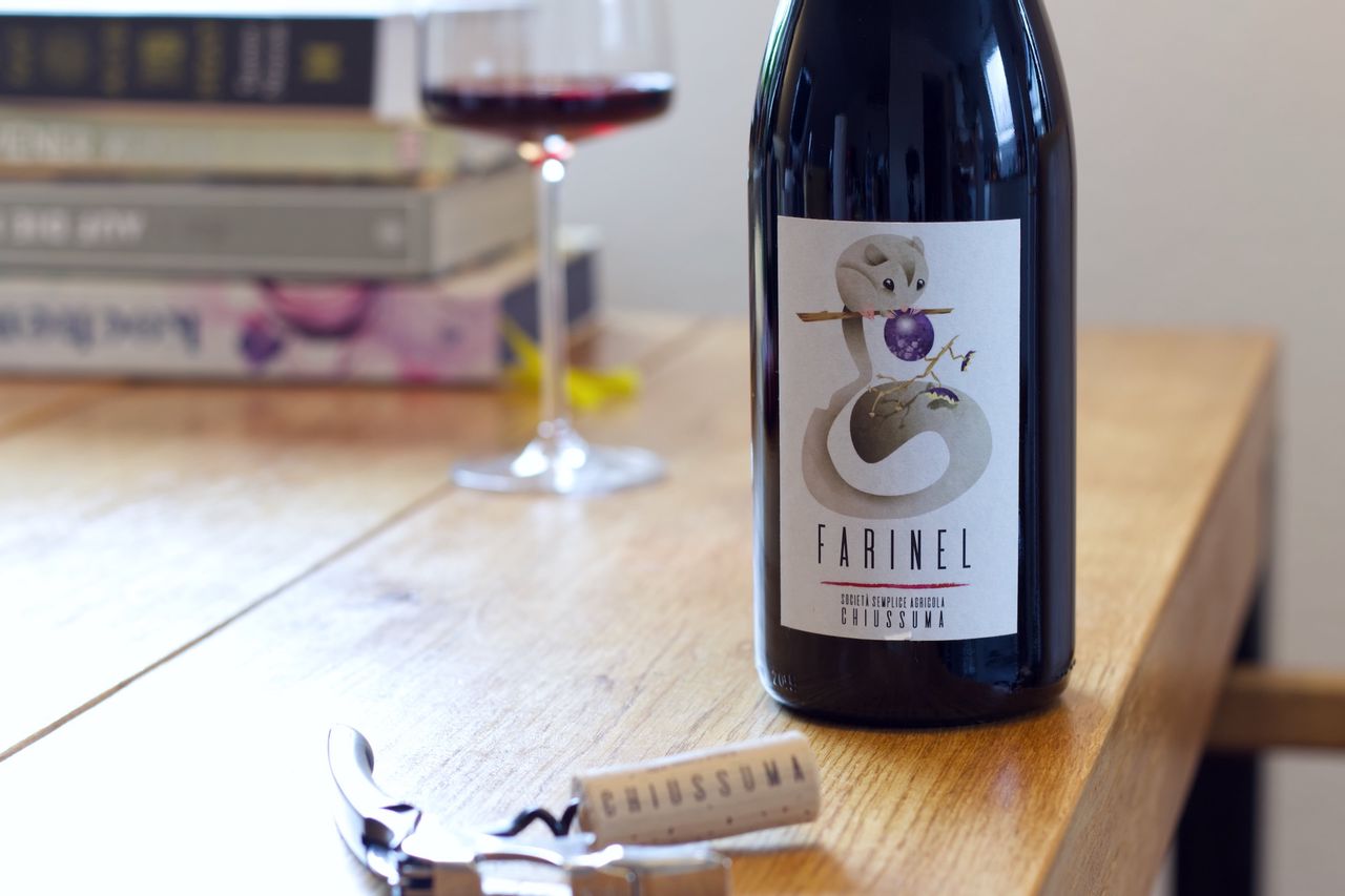 On a wooden table stands a bottle of Farinel from Chiussuma. In the background, a wine glass and a stack of books can be seen, in the foreground lies the cork on the waiter's corkscrew.