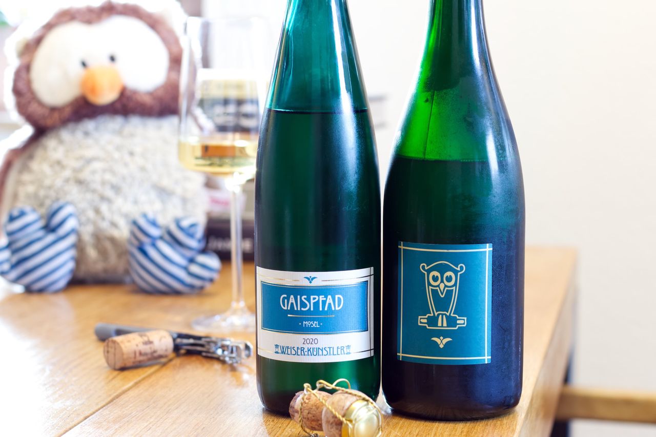 On a wooden table stand two bottles of wine from Weiser-Künstler: a Gaispfad Riesling from 2020 and a Riesling sparkling wine with an owl on the label. In the background, a wine glass, a stack of books, and an owl plush toy can be seen, in the foreground, a cork lies on the waiter's corkscrew.
