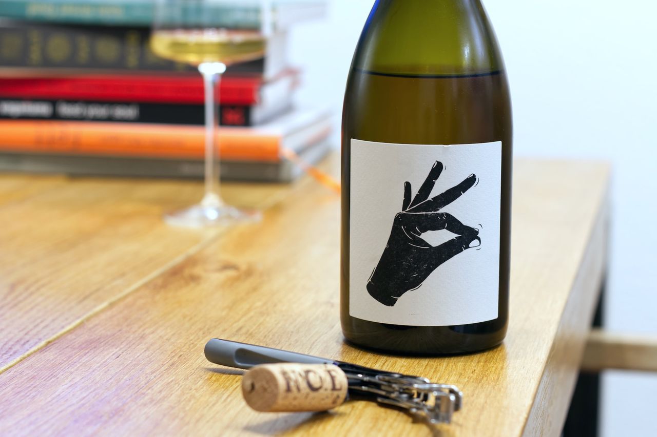 A bottle of Pattaya from the Vin Noé winery in Burgundy stands on a wooden table. The black-and-white label shows a hand rubbing two fingers together. In the background, there is a wine glass and a stack of books, in the foreground, the cork lies next to the waiter's knife.