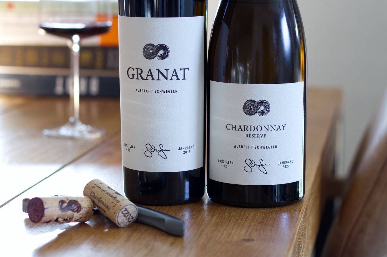 Two bottles of wine from the Albrecht Schwegler winery stand on a wooden table. In the background, a wine glass and a stack of books are visible, in the foreground lie the two corks and a waiter's knife.