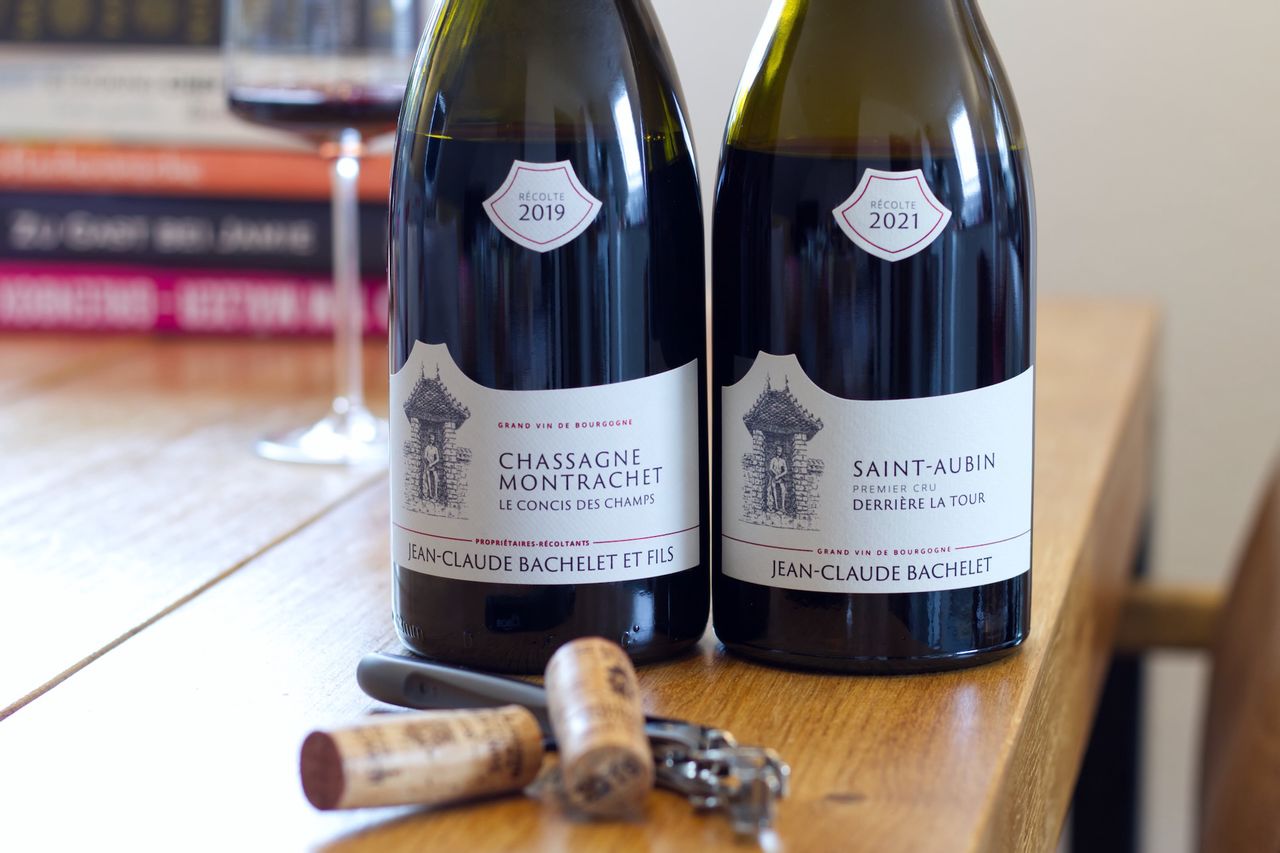 On a wooden table stand two bottles of red wine from Jean-Claude Bachelet. In the background, there is a wine glass and a stack of books; in the foreground lie the two corks and a waiter's knife.