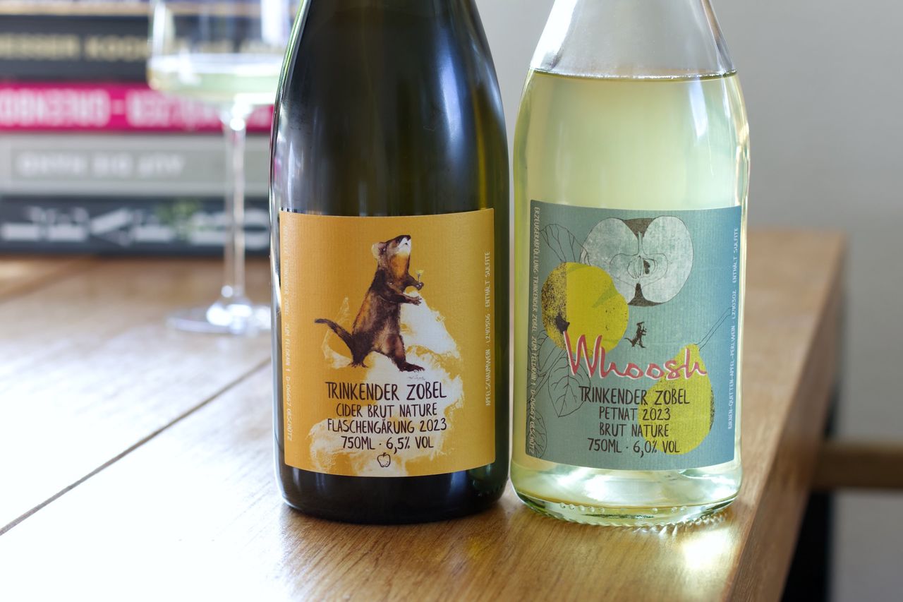 On a wooden table stand two bottles of Trinkender Zobel fruit sparkling wine. One of the labels features the namesake sable. In the background, there is a wine glass and a stack of books.