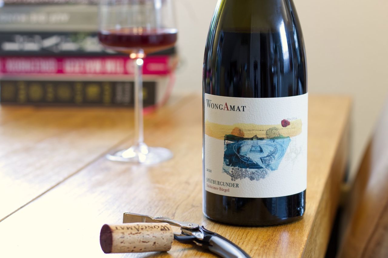 A bottle of Spätburgunder from WongAmat from the Dalsheimer Bürgel stands on a wooden table. In the background, a wine glass and a stack of books are visible, in the foreground, the cork lies next to the waiter's knife.