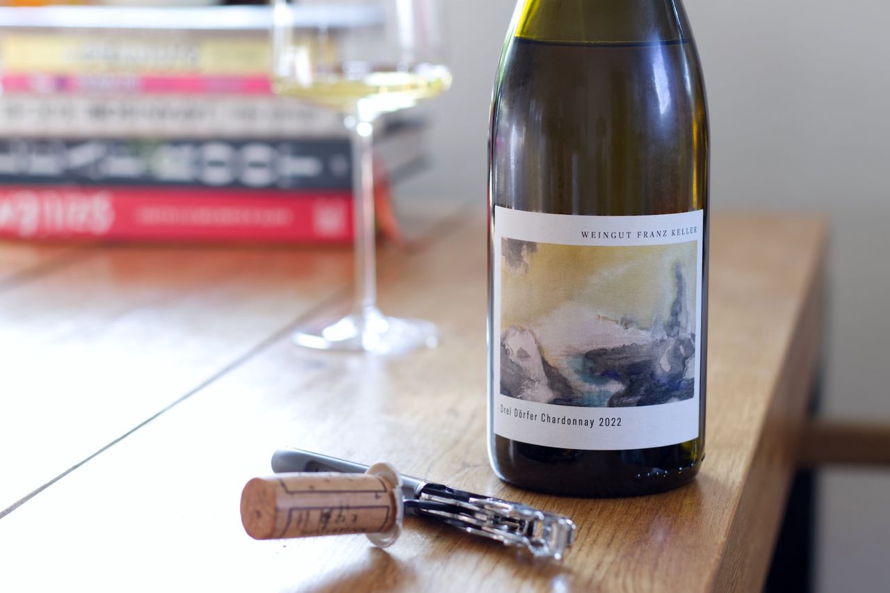 A bottle of Drei Dörfer by Franz Keller stands on a wooden table. In the background, a wine glass and a stack of books can be seen, in the foreground, the cork lies next to the waiter's knife.