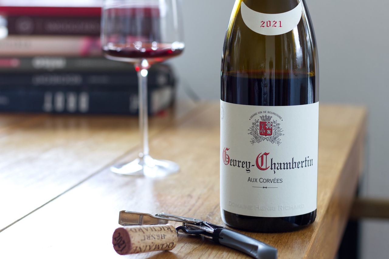 A bottle of Aux Corvées 2021 from Domaine Henri Richard stands on a wooden table. In the background, there is a wine glass and a stack of books, in the foreground, the cork rests on the waiter's knife.