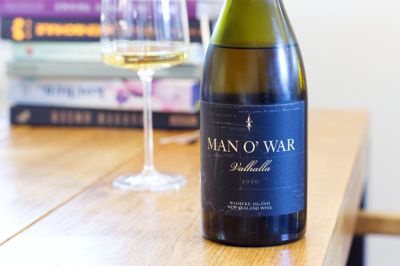 On a wooden table stands a bottle of Valhalla Chardonnay 2020 from the Man O' War winery. In the background, a wine glass and a stack of books can be seen.