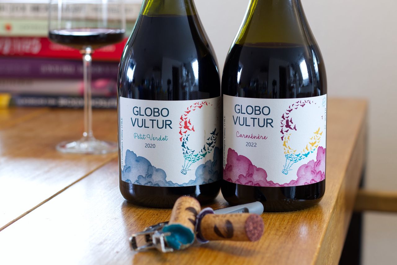On a wooden table are two bottles of red wine from Globo Vultur, a Petit Verdot and a Carménère, both featuring a hot air balloon on the label. In the background, there is a wine glass and a stack of books, in the foreground, the two corks and a waiter's knife are lying.