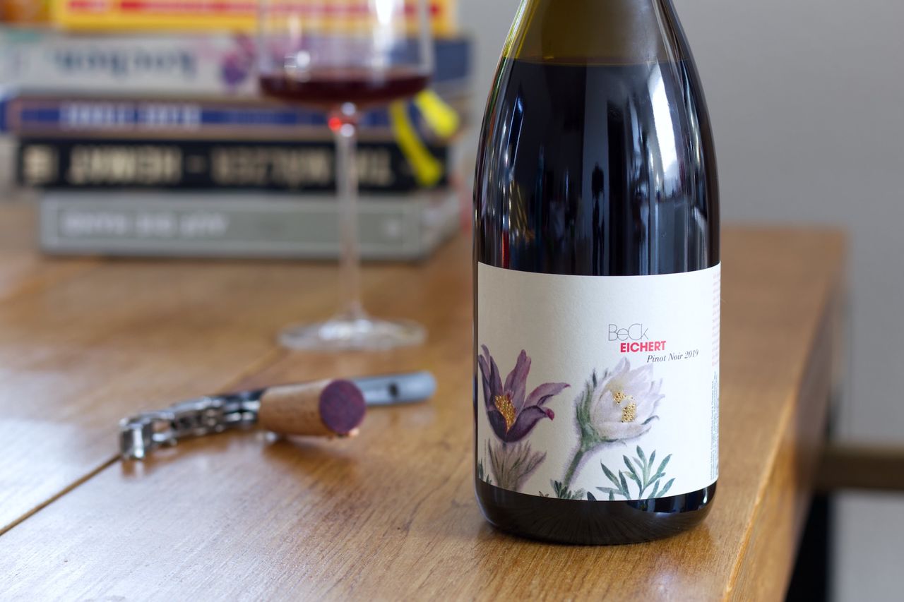 On a wooden table stands a bottle of Spätburgunder from Beck with flowers on the label. In the background, there is a wine glass and a stack of books; in the foreground, the cork lies next to the waiter's knife.