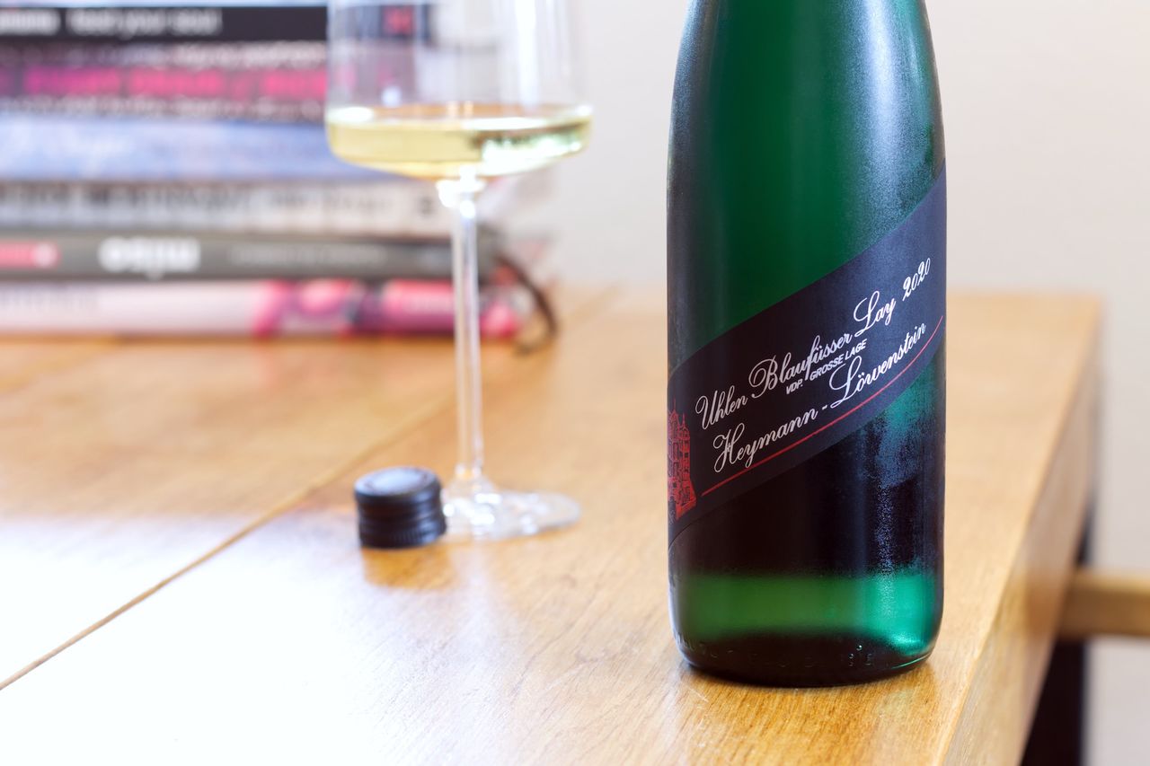On a wooden table stands a bottle of Uhlen Blaufüsser Lay 2020 Riesling from Heymann-Löwenstein. In the background, a wine glass and a stack of books are visible.