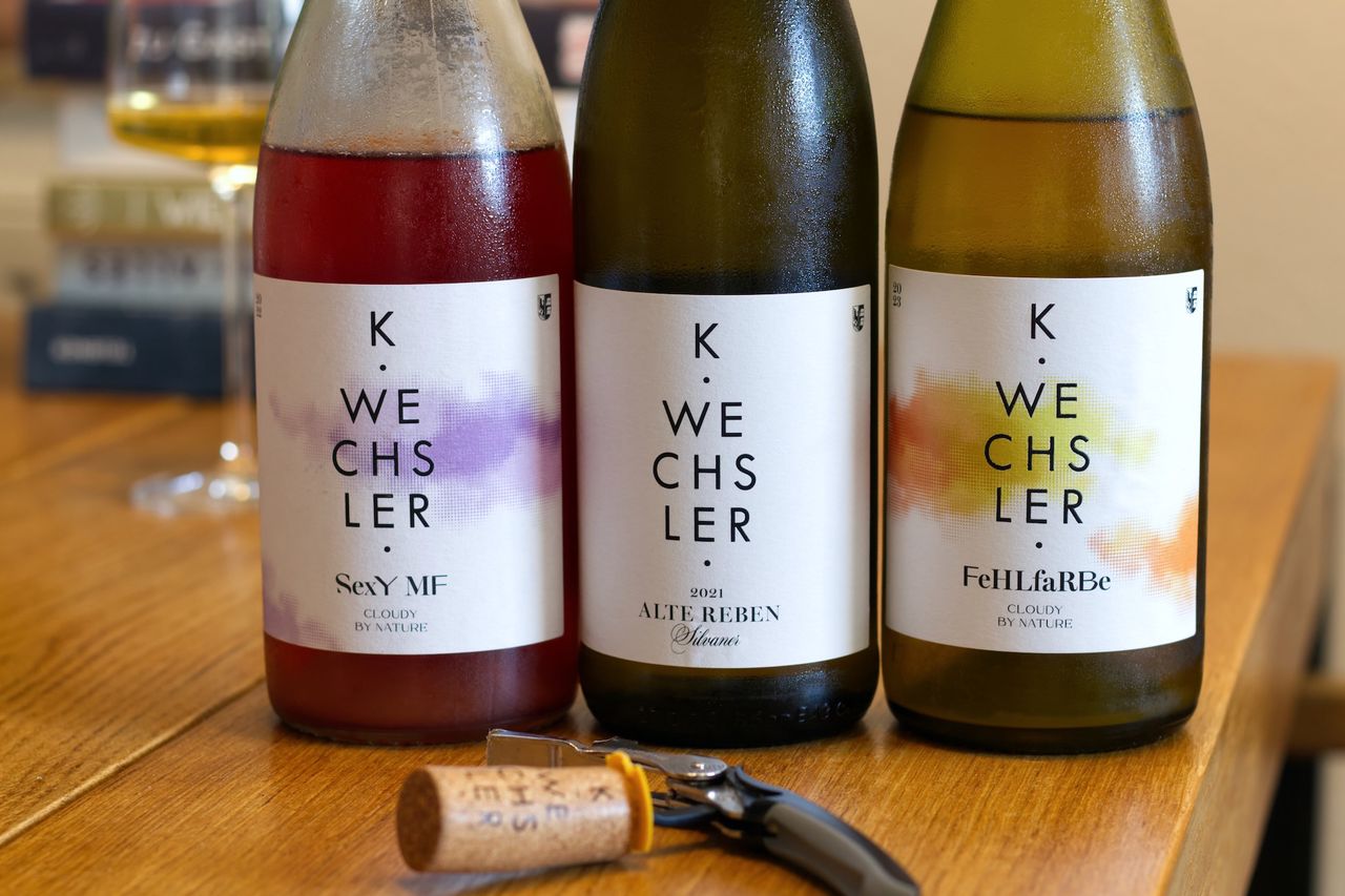 Three bottles of wine from Weingut Wechsler stand on a wooden table. The labels are simple, with black lettering and a few splashes of color. In front of them lies a cork next to a waiter's knife, behind them stands a wine glass and a stack of books.