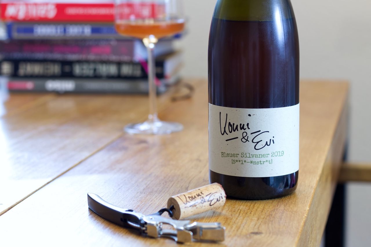 A bottle of Silvaner by Konni und Evi stands on a wooden table. In the foreground, the bottle's cork lies next to the waiter's knife. Behind the bottle, there is a wine glass and a stack of books.