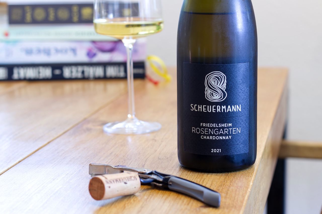A bottle of Chardonnay from the Rosengarten vineyard of Weingut Scheuermann stands on a wooden table. In the foreground, the cork from the bottle lies next to the waiter's knife. Behind the bottle are a wine glass and a stack of books.