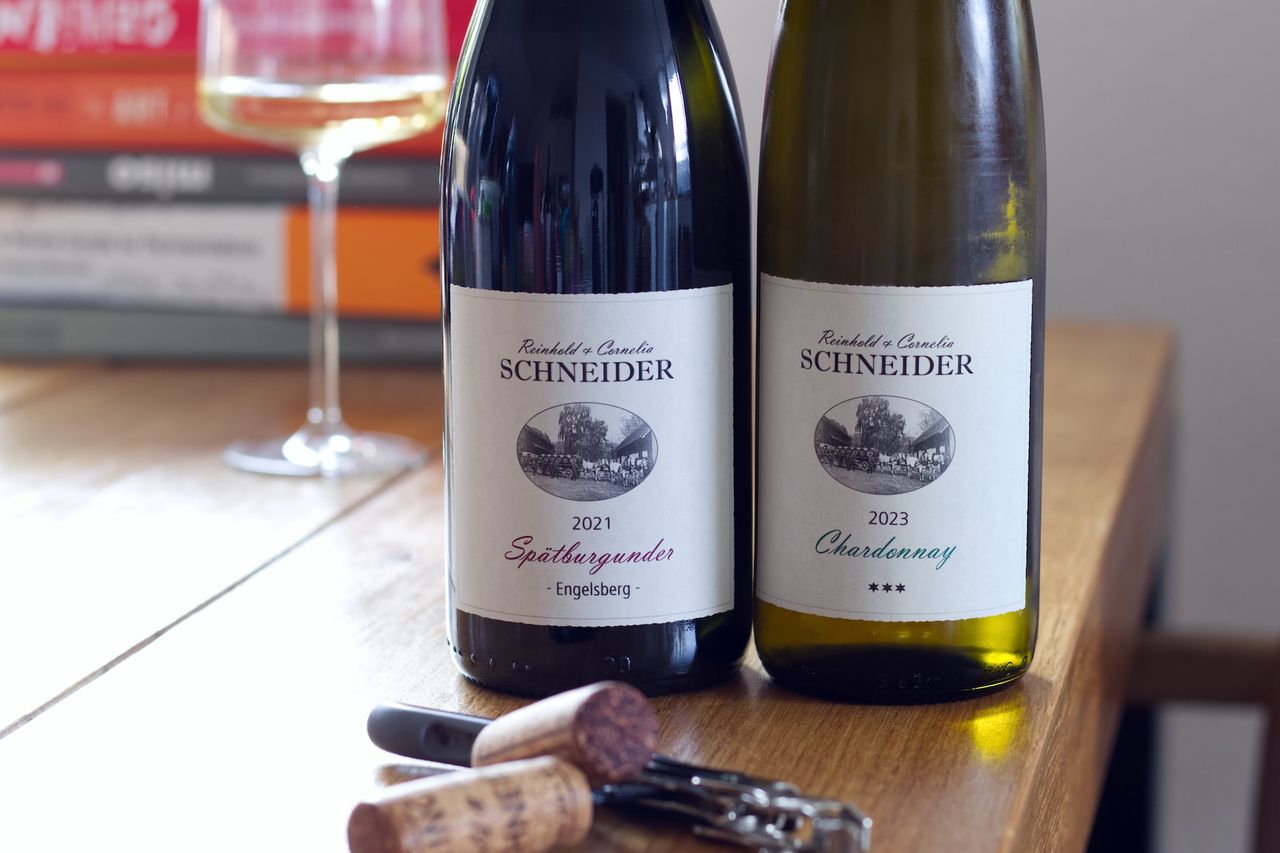 Two bottles of wine from the Reinhold and Cornelia Schneider winery stand on a wooden table. In the foreground lie the two corks and a waiter's knife. Behind them are a wine glass and a stack of books.