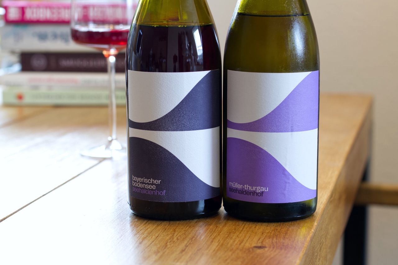 Two bottles of wine from the Seehaldenhof winery stand on a wooden table. Behind them are a wine glass and a stack of books.