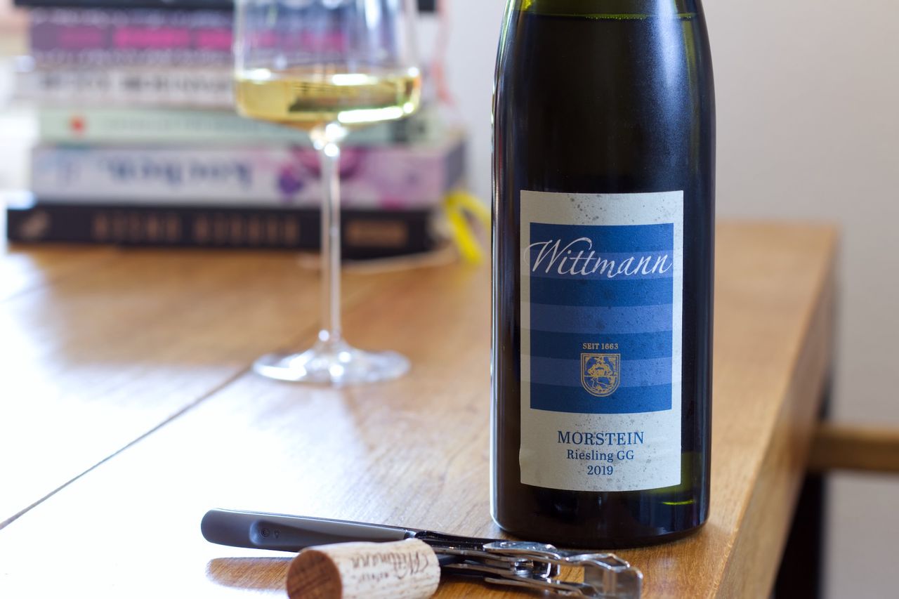 On a wooden table stands a bottle of Wittmann Riesling with the blue and white label. In the background, there is a wine glass and a stack of books; in the foreground, the cork lies next to the waiter's knife.