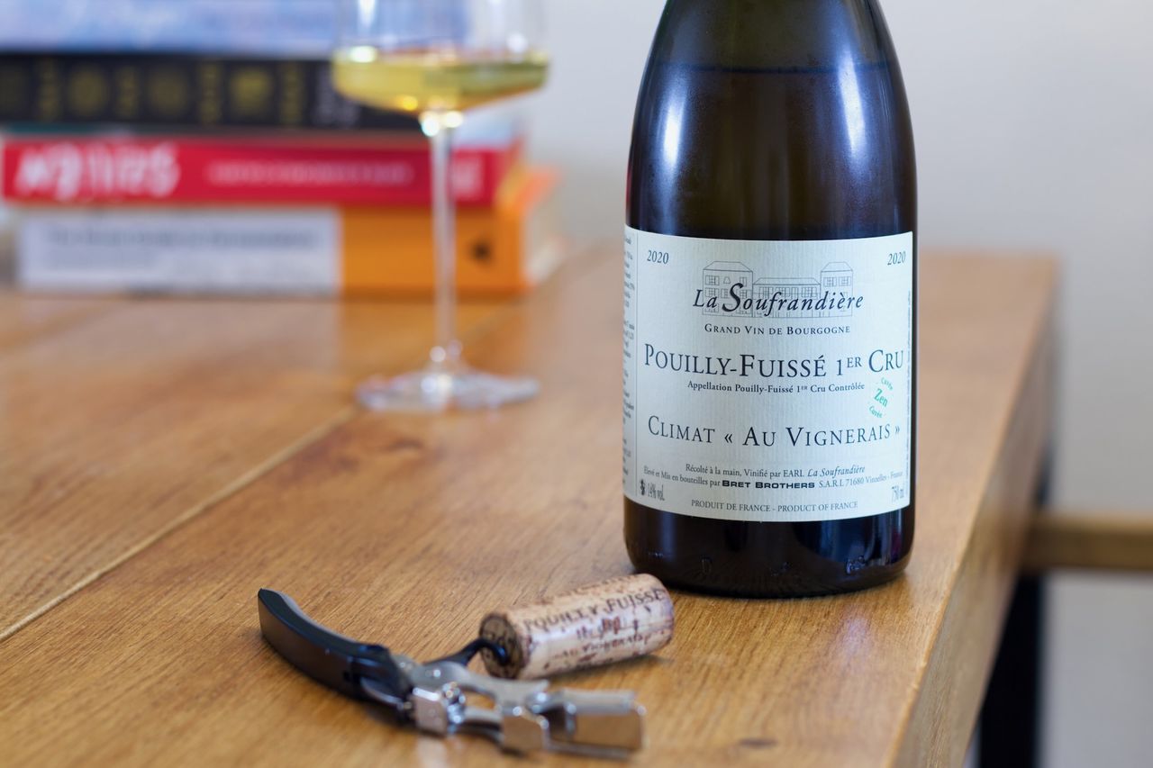 A bottle of Chardonnay Au Vignerais with a small green Zen stamp from La Soufrandière stands on a wooden table. In the foreground, the cork rests beside the waiter's knife. Behind it are a wine glass and a stack of books.