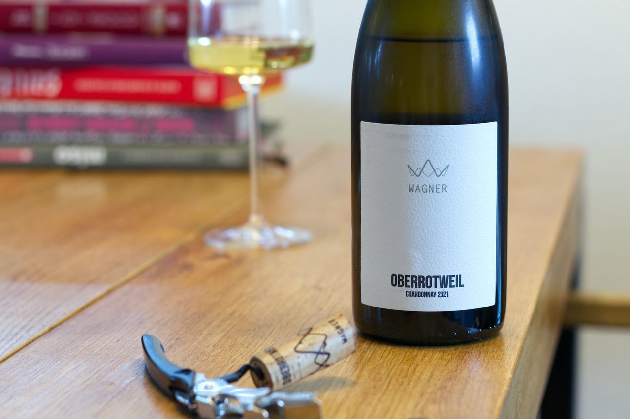 On a wooden table stands a bottle of Chardonnay from Weingut Peter Wagner. In the background, there is a wine glass and a stack of books. In front of the bottle, the cork lies next to the waiter's knife.