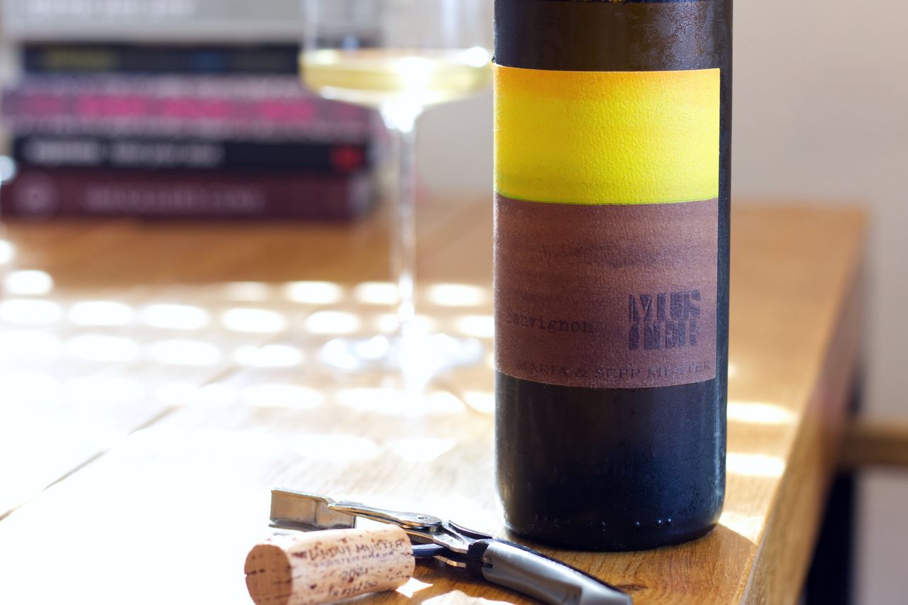 A bottle of Sauvignon 2021 from Maria and Sepp Muster stands on a wooden table. In the foreground, the bottle's cork lies on the waiter's corkscrew. Behind the bottle stand a wine glass and a stack of books.