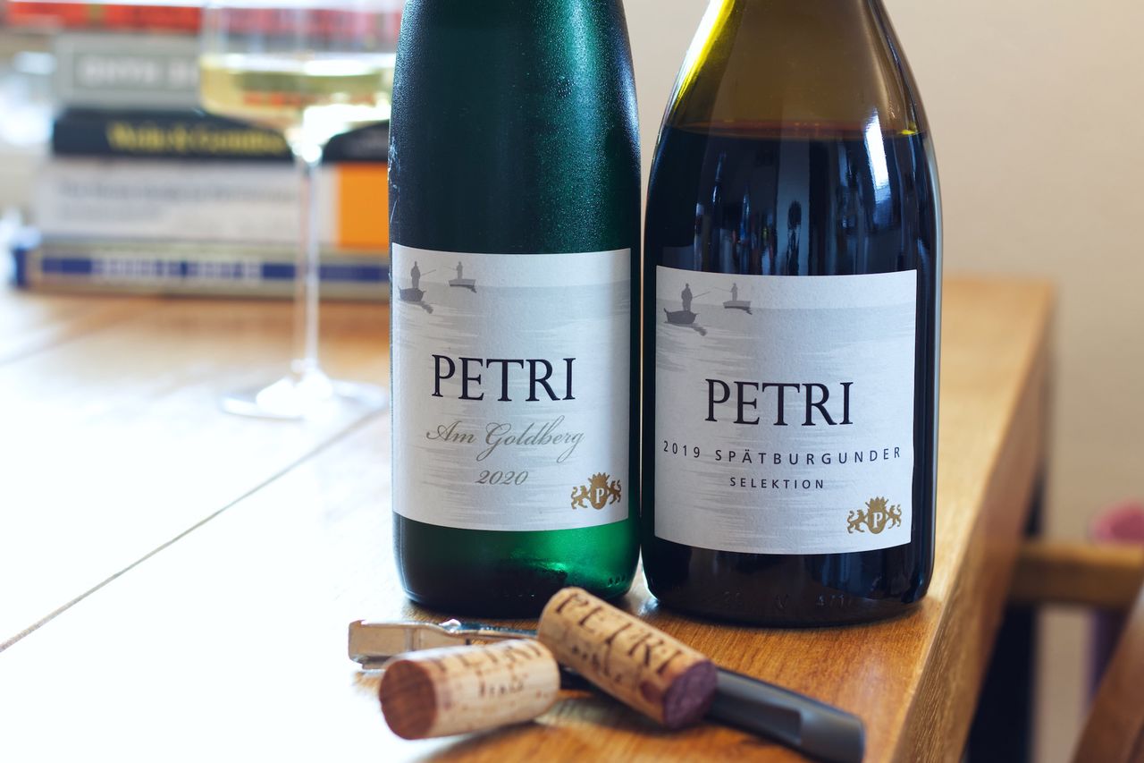 Two bottles of wine from Weingut Petri stand on a wooden table. The labels depict anglers. Behind them are a wine glass and a stack of books. In front of the bottles lie the two corks and a waiter's knife.