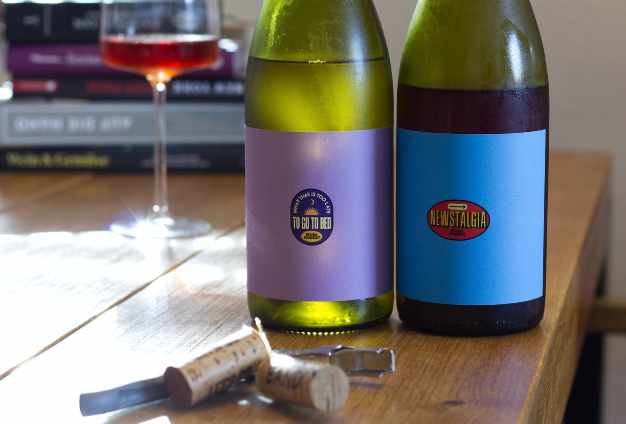 On a wooden table stand two bottles of Goutte wine. The labels are both minimalistic and striking, in vivid colors with a small logo. In the background, a wine glass and a stack of books can be seen. In front of the bottles lie corks and a waiter's knife.