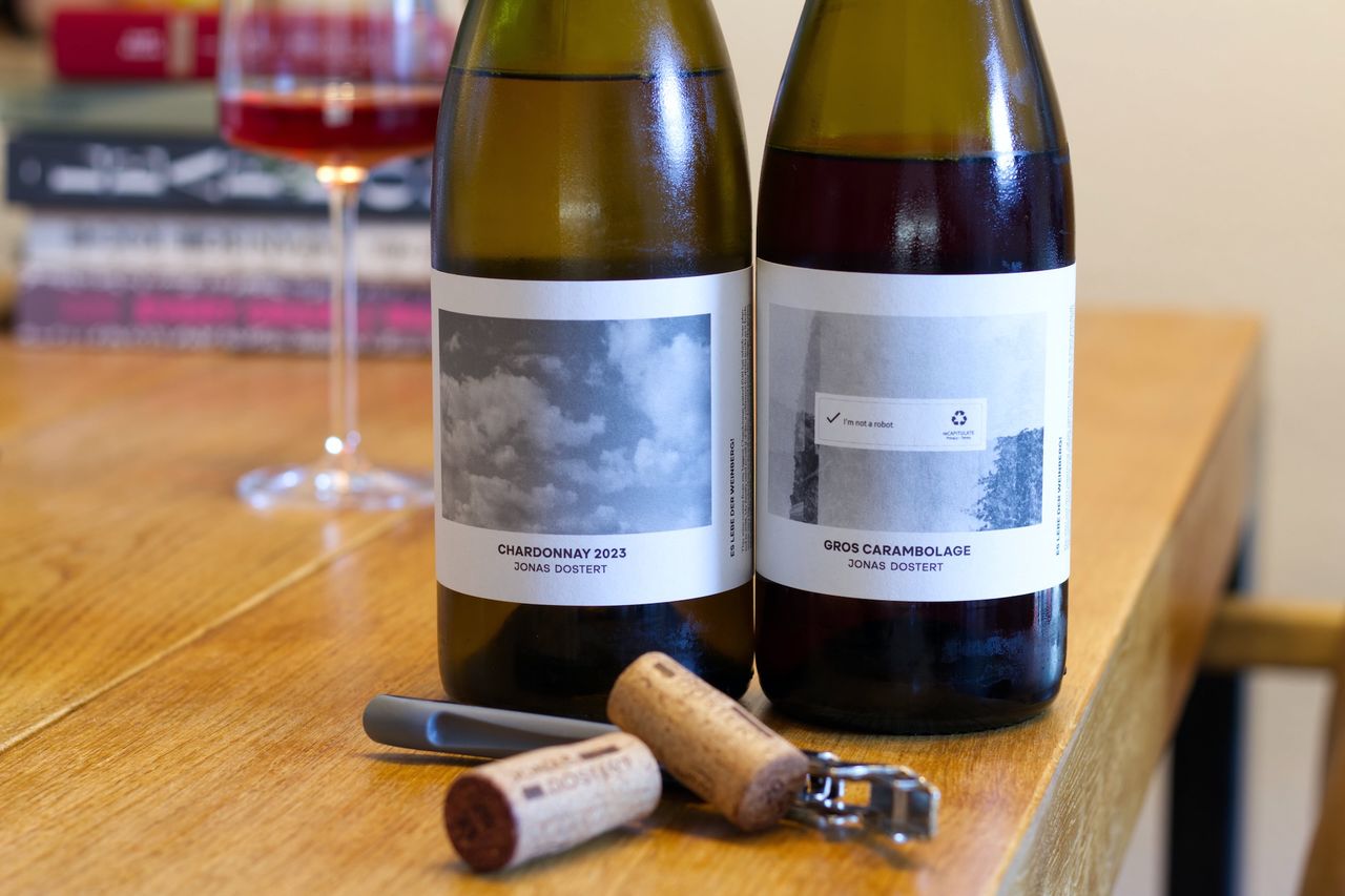 On a wooden table stand two bottles of wine from Jonas Dostert. In the background, there is a wine glass and a stack of books. In front of the bottles lie corks and a waiter's knife.