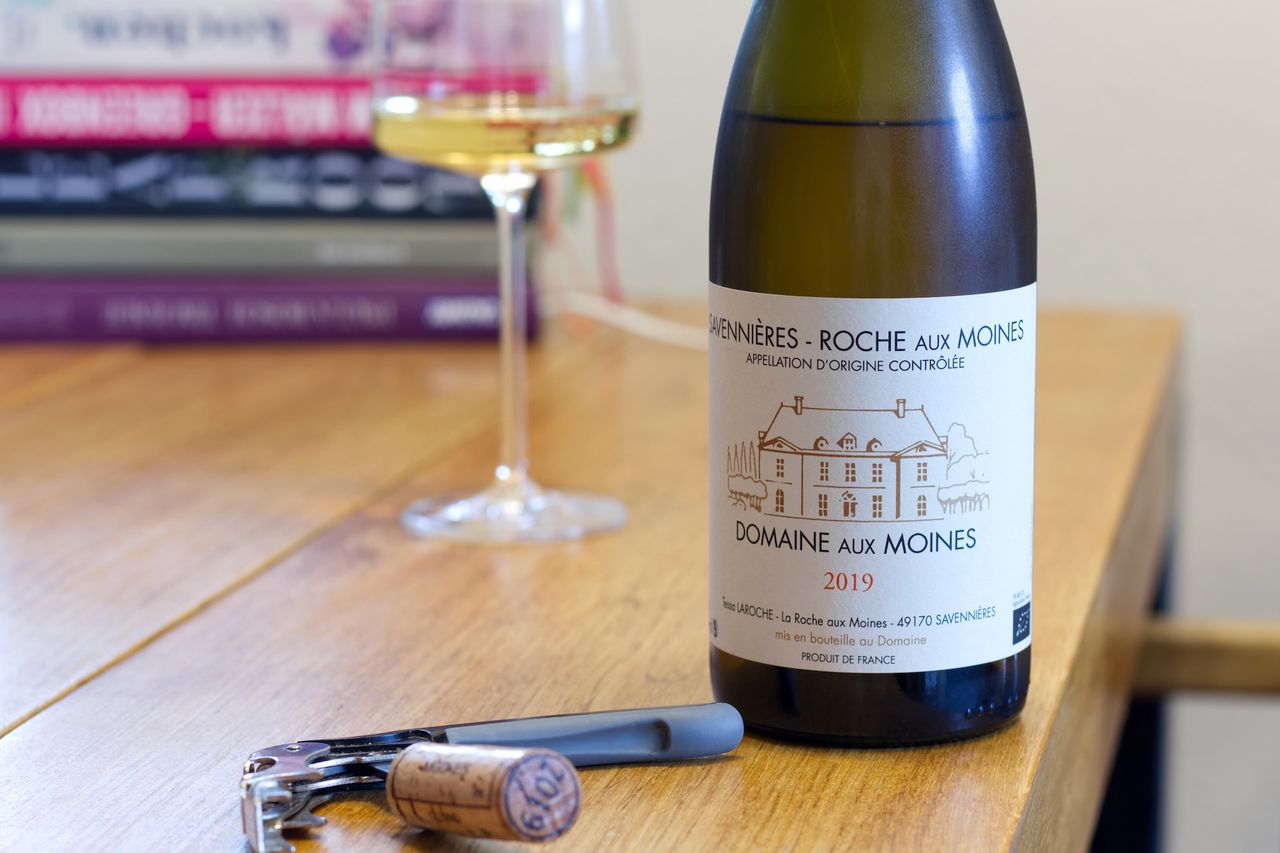 A bottle of Roche Aux Moines Chenin Blanc 2019 from Domaine Aux Moines stands on a wooden table. Behind it are a wine glass and a stack of books. In front of the bottle are the cork and a corkscrew.
