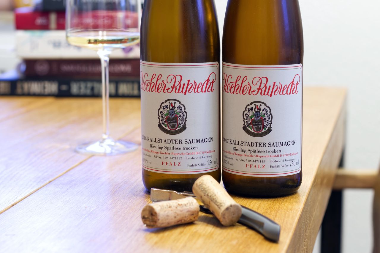 On a wooden table stand two bottles of wine from Köhler-Ruprecht. In the background, a wine glass and a stack of books are visible. In front of the bottles lie corks and waiter's knives.