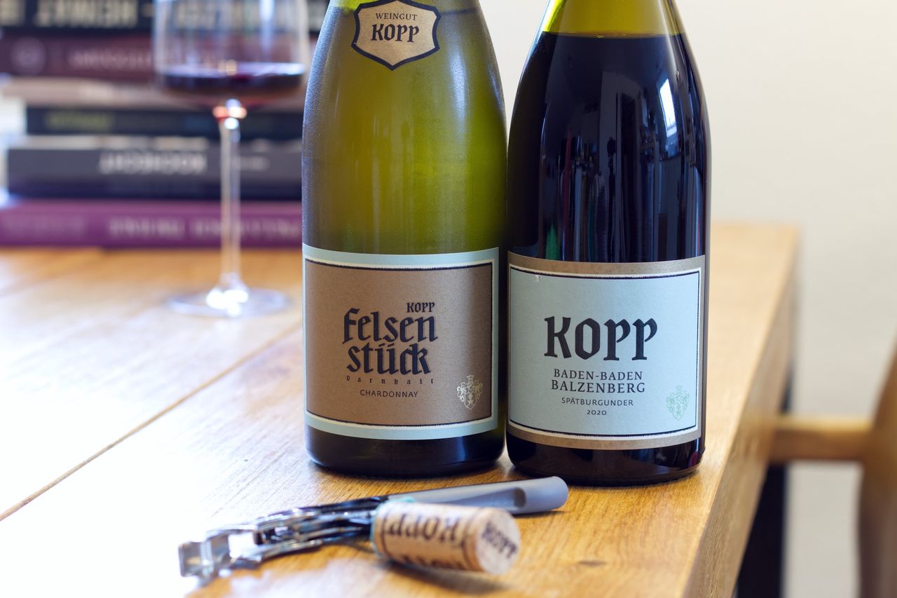 On a wooden table stand two bottles of wine from Weingut Kopp. In the background, a wine glass and a stack of books are visible. In front of the bottle lie corks and a corkscrew.