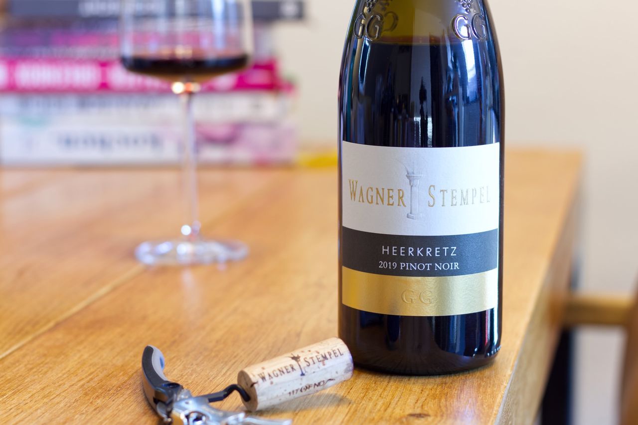 A bottle of Pinot Noir from the Heerkretz by Wagner-Stempel stands on a wooden table. In the background, there is a wine glass and a stack of books. In front of the bottle, the cork rests next to the waiter's knife.