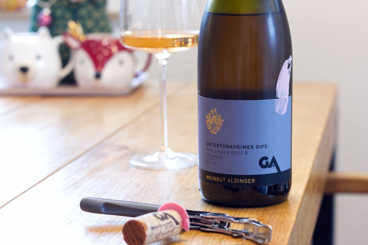 A bottle of Aldinger Trollinger Rosé with a pink brushstroke on the label stands on a wooden table. Behind it are a wine glass and a stack of books. In front of the bottles lie the cork and a Kellnermesser.