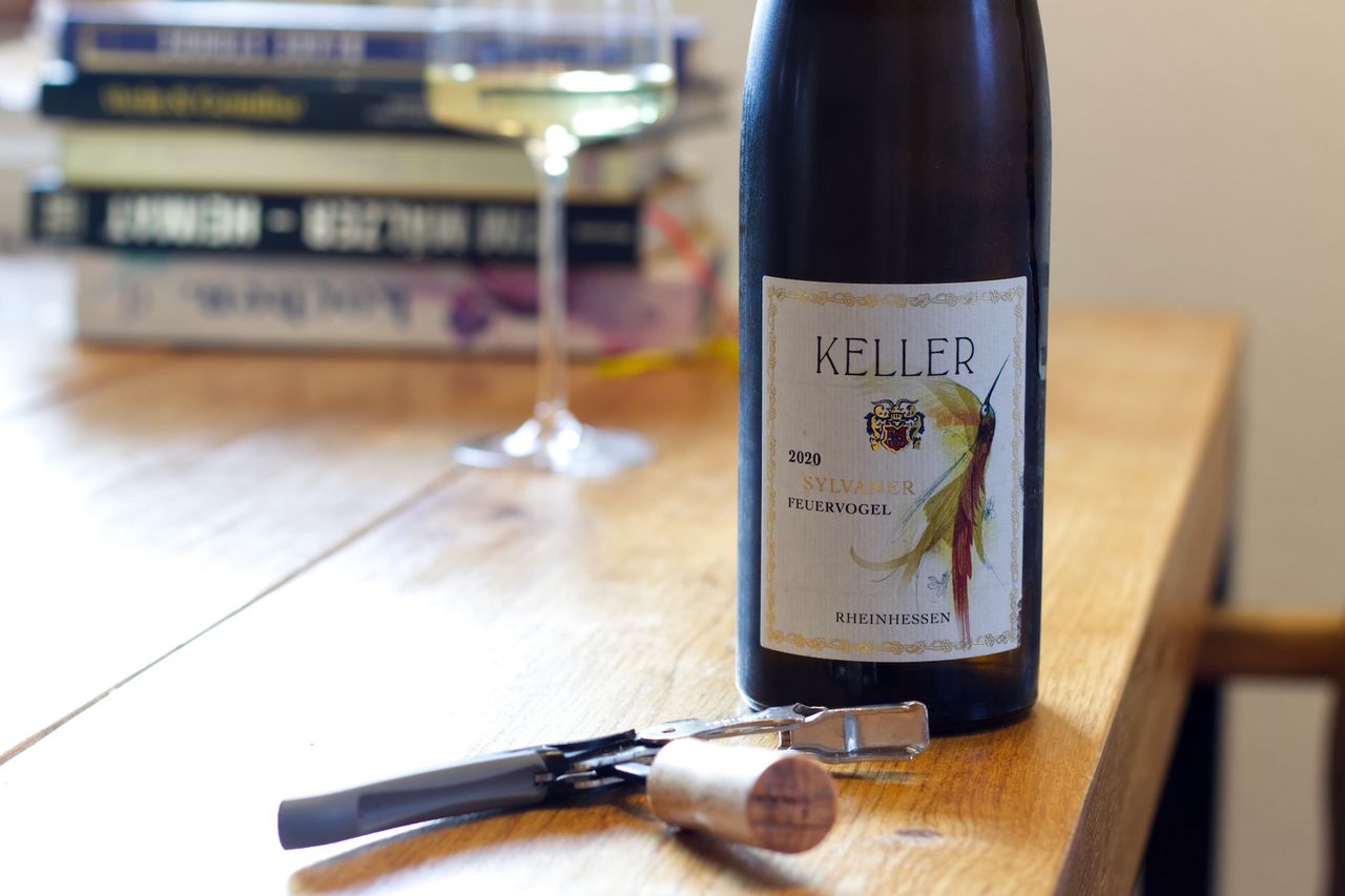 A bottle of Feuervogel 2020 from Weingut Keller stands on a wooden table. Behind it are a wine glass and a stack of books. In front of the bottles lie the cork and a Kellnermesser.