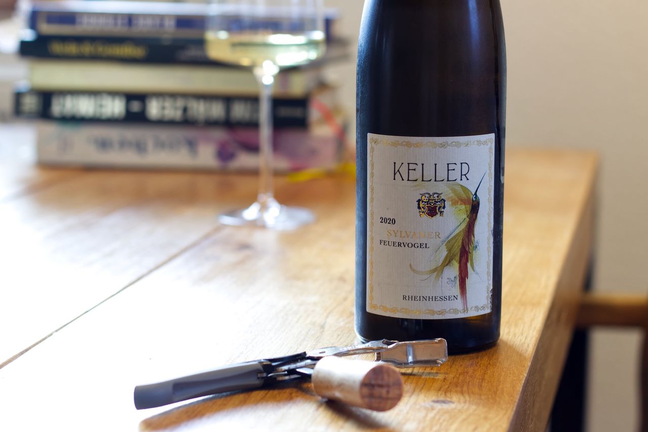 A bottle of Feuervogel 2020 from Weingut Keller stands on a wooden table. Behind it are a wine glass and a stack of books. In front of the bottles lie the cork and a Kellnermesser.