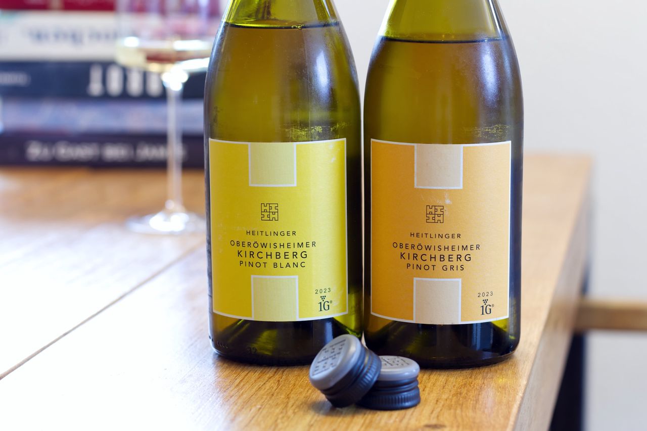 On a wooden table stand two bottles of wine from Heitlinger Winery. In the background, a wine glass and a stack of books are visible. In front of the bottles lie the screw caps.