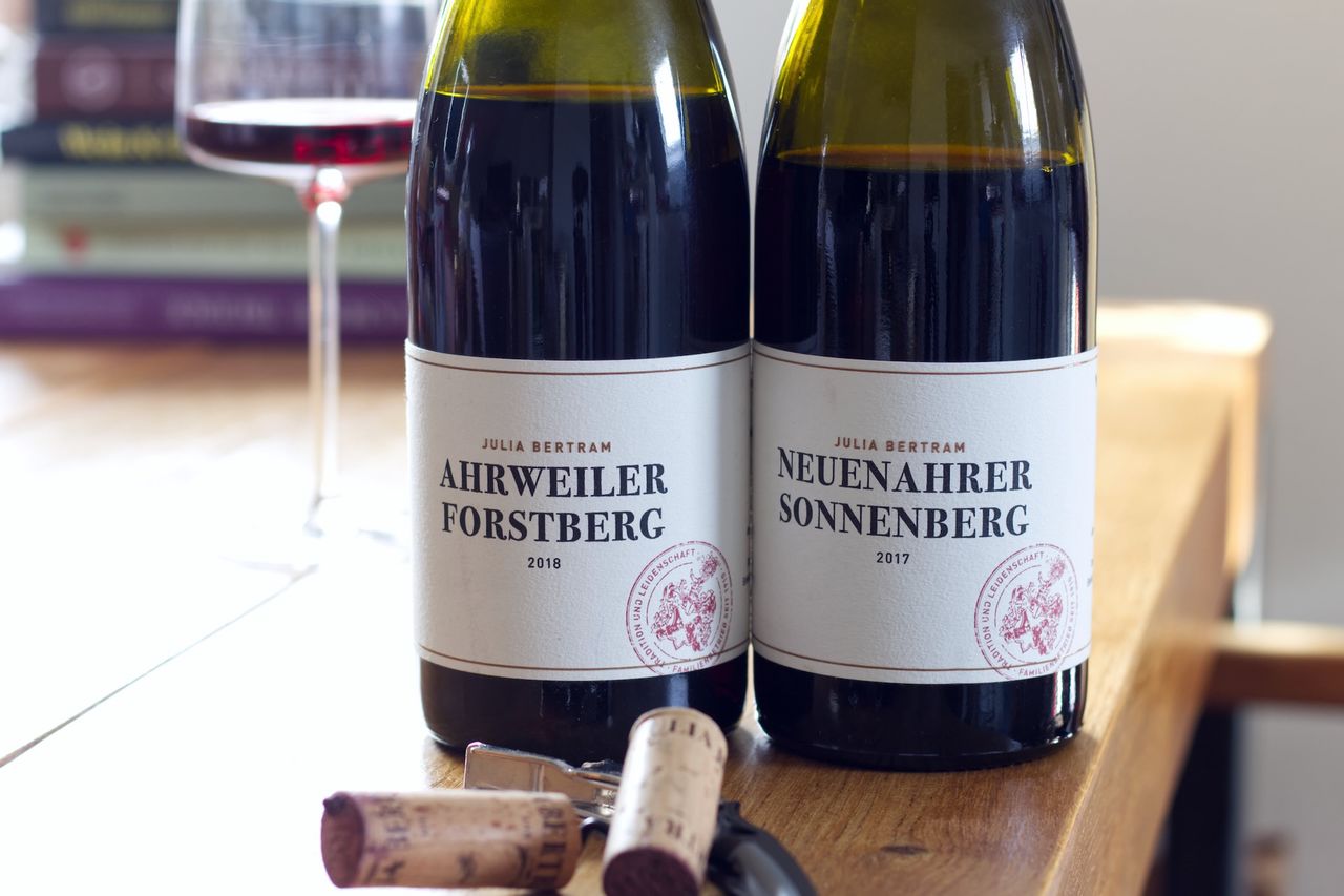 Two bottles of wine from Julia Bertram are on a wooden table. In the background, a wine glass and a stack of books are visible. Corks and a sommelier knife are lying in front of the bottle.