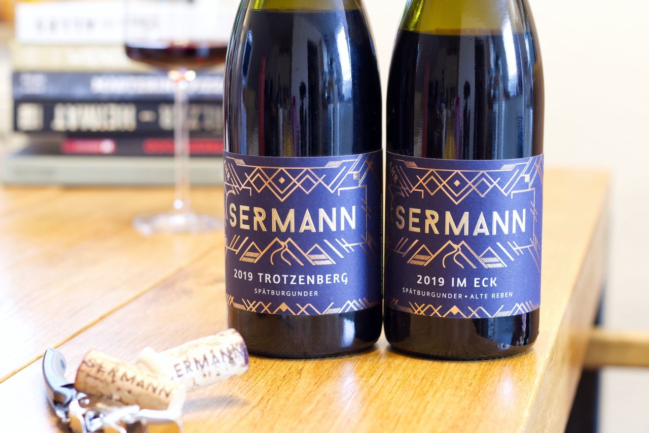 Two bottles of wine from Weingut Sermann with blue labels stand on a wooden table. Behind them are a wine glass and a stack of books. In front of the bottles lie the two corks and a waiters knife.