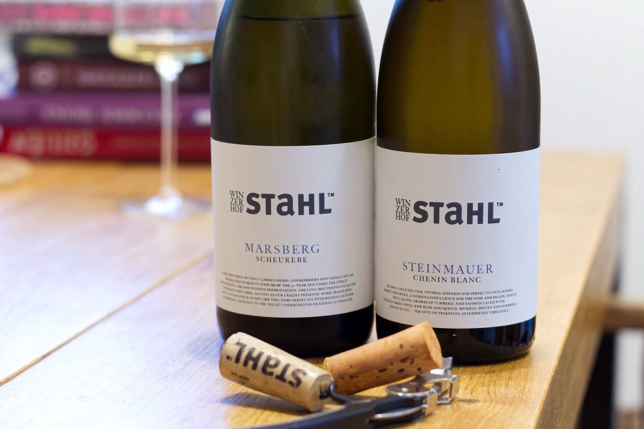On a wooden table stand two bottles of wine from Winzerhof Stahl in Franken. In the background a wine glass and a stack of books can be seen. In front of the bottles lie corks and a Kellnermesser.