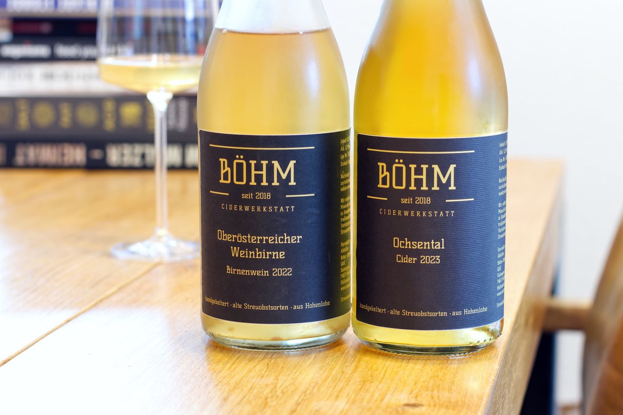 Two bottles of fruit sparkling wine from the Böhm Coderwerkstatt are standing on a wooden table. In the background, a wine glass and a stack of books can be seen.