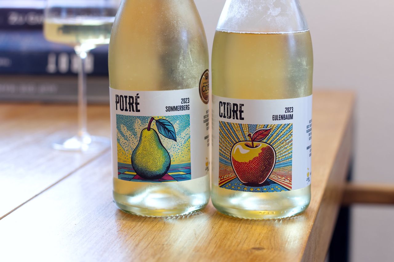 On a wooden table stand two bottles of fruit sparkling wine from Der Kleine Cidre.Betrieb. In the background, a wine glass and a stack of books can be seen.