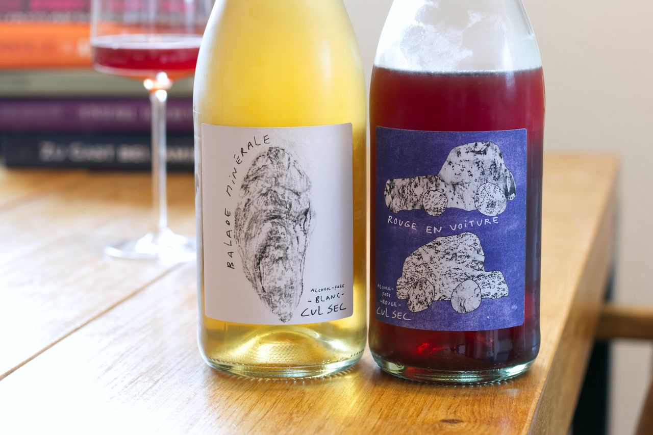 Two bottles of a sparkling wine alternative from Cul Sec stand on a wooden table. In the background, a wine glass and a stack of books are visible.