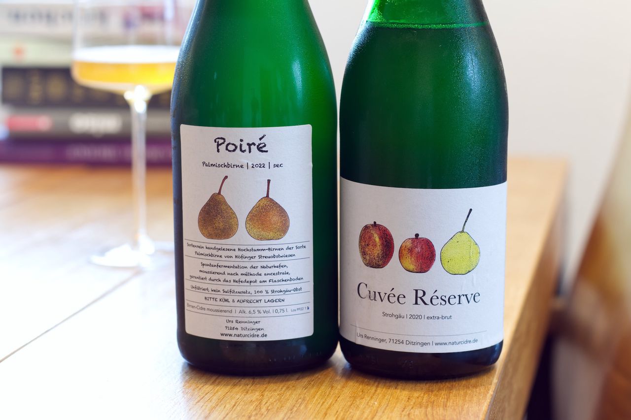 Two bottles of Obstschaumwein from Naturcidre stand on a wooden table. In the background, a wine glass and a stack of books are visible.