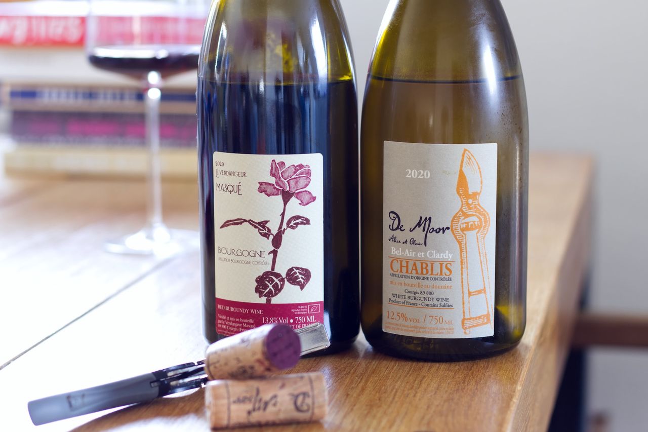 Two bottles of wine from Weingut De Moor stand on a wooden table. In the background a wineglass and a stack of books are visible. In front of the bottles, the corks lie by the waiter's knife.