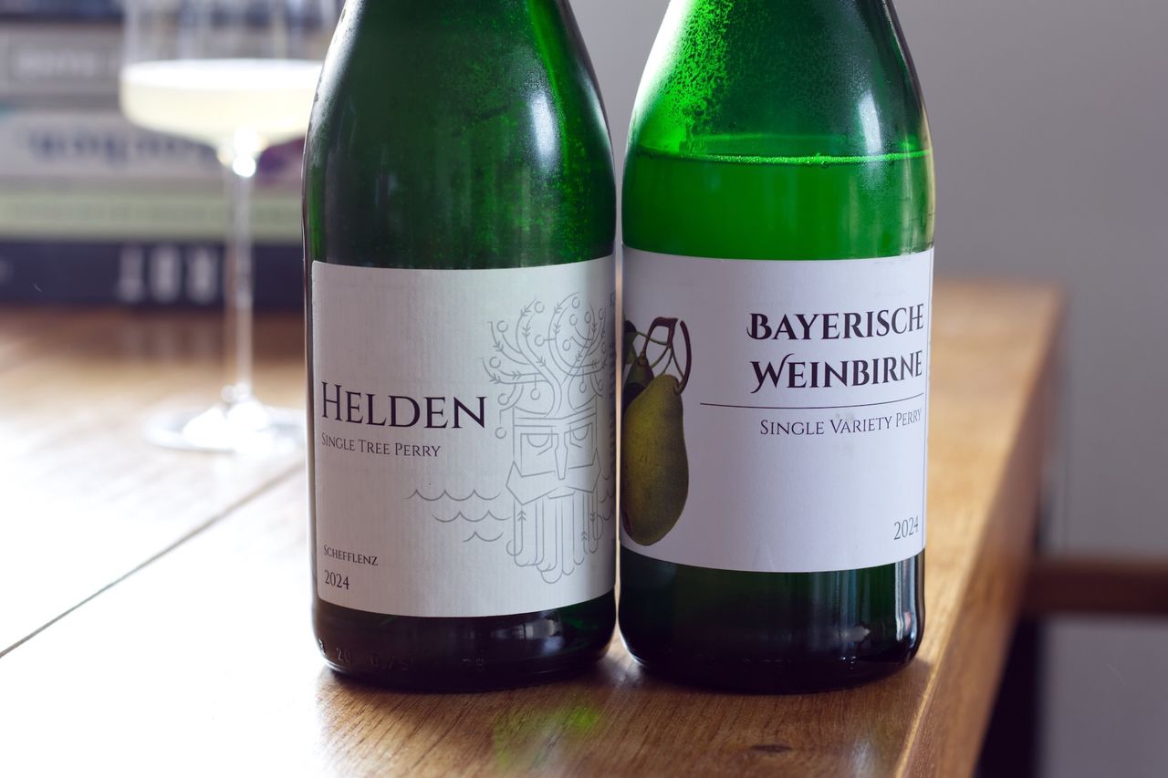 Two bottles of Kertelreiter sparkling fruit wine stand on a wooden table. In the background, a wine glass and a stack of books are visible.