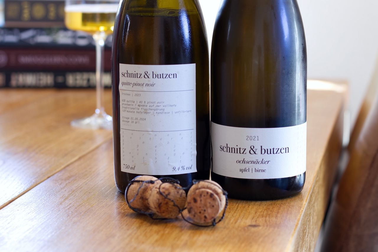 Two bottles of fruit sparkling wine from Schnitz and Butzen stand on a wooden table. In the background, a wine glass and a stack of books are visible.