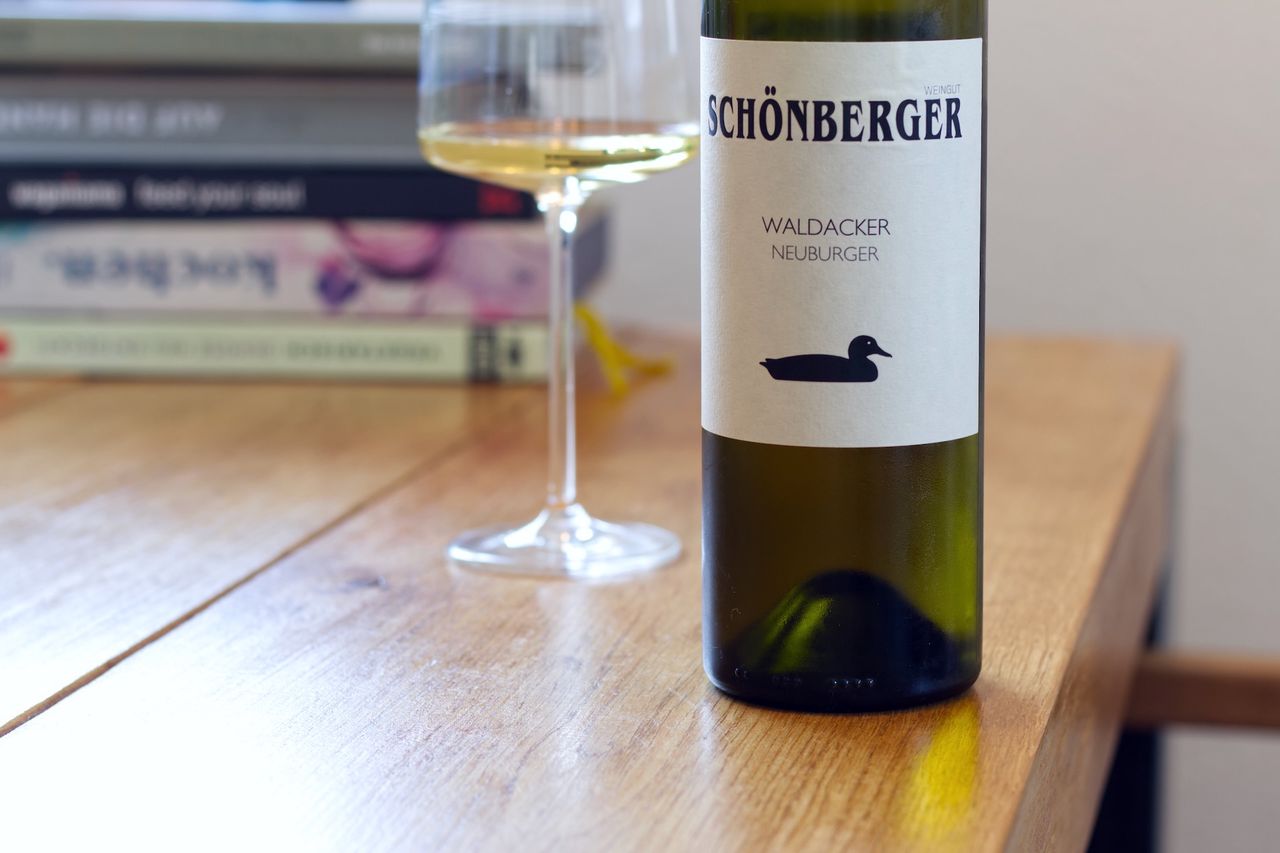 A bottle of Neuburger from Schönberger with the duck on the label stands on a wooden table. In the background, a wine glass and a stack of books are visible.
